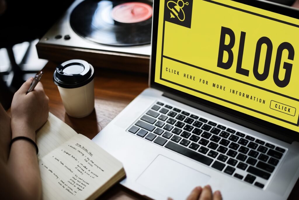 Seorang penulis membuat catatan di samping laptop dengan tampilan layar bertuliskan BLOG, menggambarkan aktivitas menulis dan jurnalisme digital.